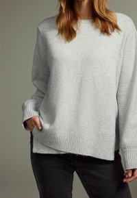 Pull en maille gris clair avec un col rond, des poignets et un ourlet côtelés, et une coupe décontractée. Tissu texturé et fentes latérales visibles.