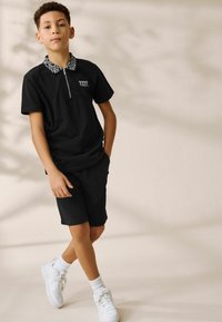 Polo nero con colletto a motivo, collo con zip, abbinato a pantaloni corti neri. Indossato con scarpe da ginnastica bianche e calzini alla caviglia. Tessuto morbido.