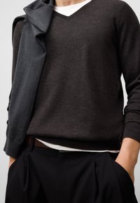 Brauner V-Ausschnitt-Pullover aus feinem Strickmaterial, kombiniert mit einem dunkelgrauen Wickelmantel. Übereinander getragen mit einem weißen Hemd und schwarzen Hosen mit Falten.