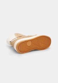 Zapatilla con parte superior de ante marrón, acentos crema, suela blanca y suela exterior de goma texturizada con surcos y patrones circulares.