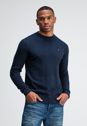 Maritimer Strickpullover mit strukturiertem Muster, Rundhalsausschnitt und gerippten Bündchen. Schwarzes Logodetail auf der Brust. Kombiniert mit blauen Jeans.