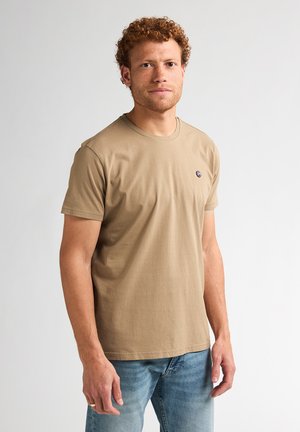 Man met krullend rood haar, gekleed in een beige T-shirt met korte mouwen en lichtblauwe jeans, staand tegen een eenvoudige lichte achtergrond.