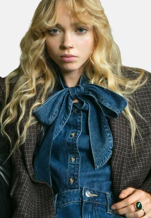 Koszula dżinsowa z dużym kołnierzem w kształcie kokardy, zestawiona z pikowaną brązową marynarką. Posiada złote guziki i zielony pierścionek na dłoni.