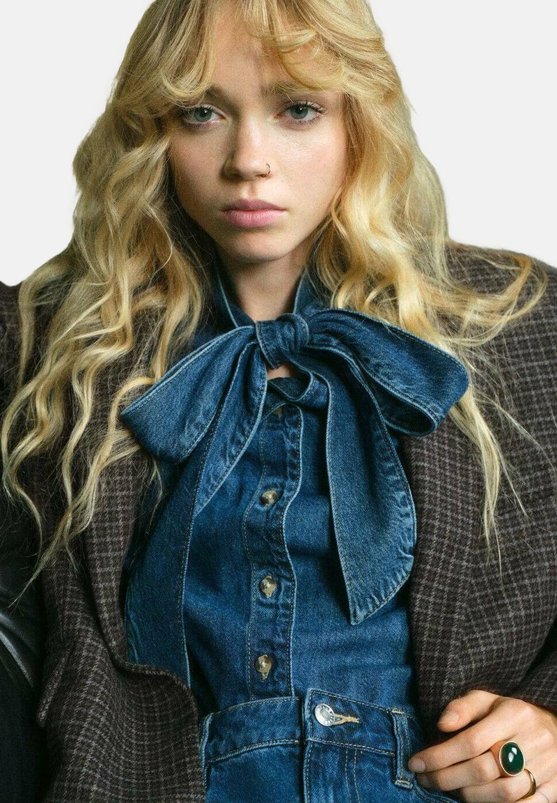 Koszula dżinsowa z dużym kołnierzem w kształcie kokardy, zestawiona z pikowaną brązową marynarką. Posiada złote guziki i zielony pierścionek na dłoni.