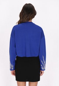 Rövidített kék pulóver hosszú ujjakkal, ezüst hímzett díszítéssel az ujjak végén, fekete szoknyával párosítva. Sima textúra.