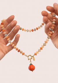 Collar de cuentas que presenta cuentas en tonos naranja, durazno y gris con acentos dorados. Un gran charm rojo en el cierre añade un detalle notable.