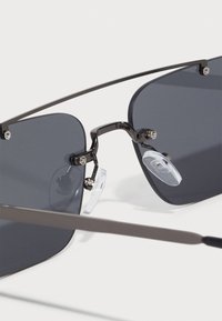 Kovové sluneční brýle se šedými skly, tenkým rámem a průhlednými nosními podložkami. Disponují minimalistickým designem a elegantními liniemi.