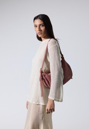 Frau mit langen dunklen Haaren, die einen cremefarbenen Pullover und einen beigen Rock trägt, trägt eine staubig-rosafarbene Lederschultertasche vor einem schlichten Hintergrund.