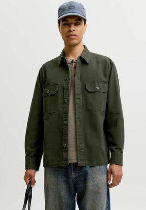 Homme portant une casquette grise, une chemise verte olive boutonnée sur un henley beige, un jean bleu, tenant une anse de sac noire sur un fond uni.