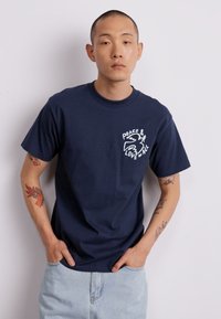 T-shirt de algodão azul marinho com mangas curtas, apresentando texto e gráfico brancos "PAZ & AMOR A TODOS" no peito. Ajuste casual, decote redondo padrão.