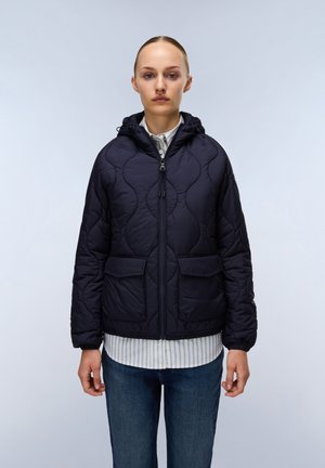 Veste matelassée bleu marine avec capuche, fermeture éclair et deux poches avant. Présente un motif texturé et des poignets élastiqués. Portée sur une chemise rayée.
