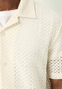 Camisa de manga corta en color crema con un patrón de encaje. Presenta un cuello, cierre con botones y un tejido texturizado, que exhibe detalles de diseño calado.
