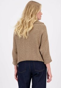 Bruine, gestructureerde trui met een kraag en driekwartmouwen, gecombineerd met donkerblauwe jeans. De stof heeft een chunky breipatroon.