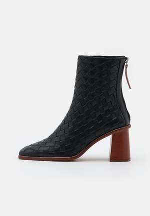 Schwarzer geflochtener Leder-Stiefelette mit spitzer Kappe, braunem Blockabsatz und Rückenzipper mit Lederlasche vor weißem Hintergrund.