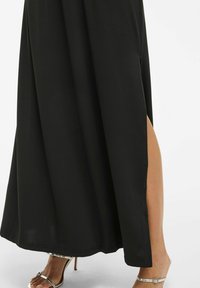 Robe maxi noire avec une fente sur le côté, tissu lisse, design long jusqu'au sol, et des escarpins ouverts argentés visibles. Silhouette simple et élégante.