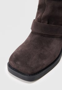 Brune suede ankelstøvlett med firkantet tå og sort gummisåle. Har en sydd søm og et bredt belte over vristen.