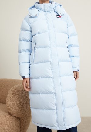 Personne portant un long manteau matelassé bleu clair avec un col montant et une capuche, debout à côté d'une chaise beige texturée.