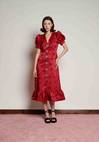 Κόκκινο φόρεμα από σατέν με φουσκωτά μανίκια, V-λαιμόκοψη, πέντε κουμπιά σε σχήμα καρδιάς, φλοράλ σχέδιο και κρόσσια στο τελείωμα. Μαύρα πέδιλα με λουριά ολοκληρώνουν την εμφάνιση.