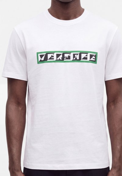 Homme portant un t-shirt blanc avec un logo rectangulaire noir bordé de vert affichant un texte blanc stylisé indiquant "VERSACE" sur la poitrine.
