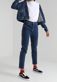 Person som bär en mörkblå denimjacka och jeans, vit t-shirt, röda strumpor och svarta sneakers med vita snören, står med en arm höjd.