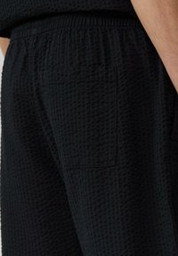 Pantalon en tissu noir à texture gaufrée, doté d'une taille élastique et d'une poche au dos. La vue en gros plan met en évidence la texture et le design de la poche.
