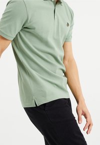 Polo shirt vert clair avec un tissu texturé, deux boutons au col, fentes latérales et un petit logo sur la poitrine, associé à un pantalon noir.