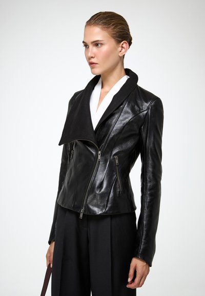 Femme portant une veste en cuir noire ajustée avec une fermeture éclair asymétrique et un large col, associée à un pantalon noir, debout devant un fond blanc.