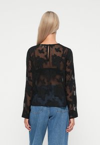 Sort gennemsigtig bluse med lagdelte blomsterapplikationer. Lange ærmer, rund hals med nøglehulslukning bagpå. Sammen med blå denim jeans.