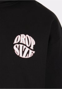 Tessuto nero con testo bianco in stile bolla "DROP SIZE" distorto in forma circolare nella zona superiore del petto.