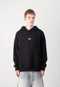 Schwarzer Kapuzenpullover aus Baumwolle mit einer Fronttasche. Verfügt über ein kleines weißes Logo-Tag im Brustbereich. Modell trägt helle Jeansshorts.