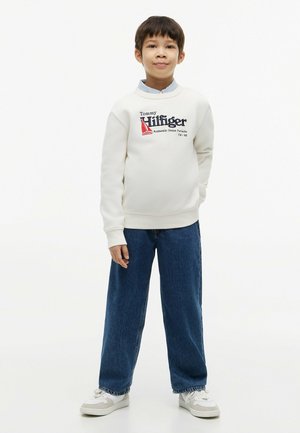 Ragazzo in piedi con le mani nelle tasche, indossando una felpa bianca Tommy Hilfiger, jeans blu e sneakers bianche su sfondo bianco.