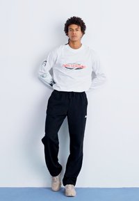 Biely dlhý rukáv s logom "new balance", čierne tepláky a béžové tenisky; model stojí na jednoduchom pozadí.