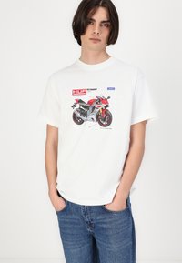 Hvid bomulds t-shirt med grafik af en rød motorcykel med sorte detaljer. Korte ærmer og afslappet pasform. Blå jeans på.