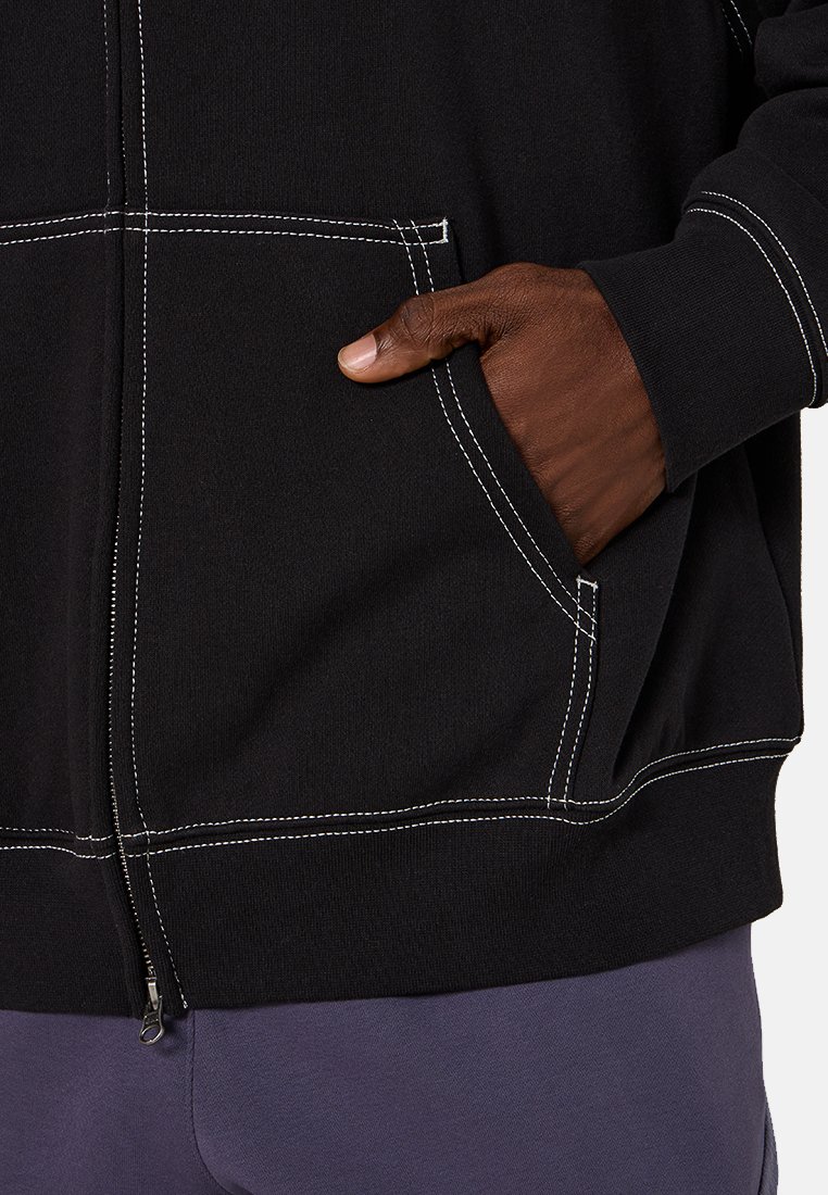 Sudadera con cremallera negra con detalles de costura blanca. Cuenta con bolsillos frontales y una textura de tela robusta. Mano del modelo posicionada en el bolsillo.