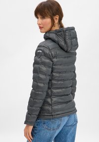 Vrouw met bruin haar, gekleed in een donkergrijze gewatteerde gevoerde jas met capuchon en blauwe spijkerbroek, iets naar de zijkant gedraaid tegen een witte achtergrond.