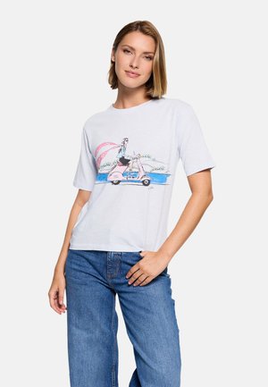 Mujer con camiseta blanca con una ilustración colorida de una persona montando un scooter rosa junto al agua, combinada con jeans azules.