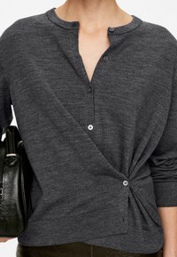 Grau-meliertes, leichtes Strickpullover mit Henley-Ausschnitt, zwei Knöpfen und einem verdrehten Saumdetail. Das Model hält eine schwarze Tasche.