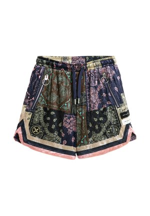 Joven de pie con camisa de manga corta con patrón de cachemira multicolor y pantalones cortos a juego, con calcetines blancos y zapatos beige texturizados sin cordones.