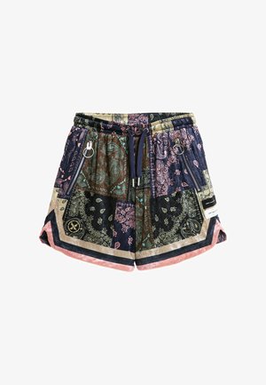 Joven de pie con camisa de manga corta con patrón de cachemira multicolor y pantalones cortos a juego, con calcetines blancos y zapatos beige texturizados sin cordones.