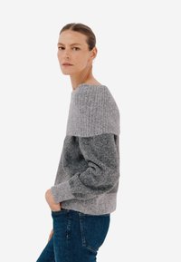 Szary sweter wykonany na drutach z ribbowanym dekoltem i teksturowanym ciałem, charakteryzujący się luźnym krojem i długimi rękawami, w zestawieniu z niebieskimi dżinsami.