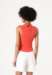 Rotes geripptes Tanktop mit taillierter Passform, das eine Bindeschleife am Halsrücken hat und mit weißen Denim-Shorts kombiniert ist.