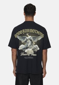 Maglietta nera in cotone con graphic di un angelo alato, dettagli in oro e bianco sul retro, il testo recita "GOODFORNOTHING" e "Heavenly Chaos."