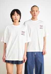 White oversized t-shirts feature a small multicolored logo on the left chest. Model pairs with denim shorts and striped blue pants.