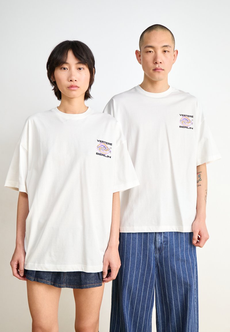 White oversized t-shirts feature a small multicolored logo on the left chest. Model pairs with denim shorts and striped blue pants.
