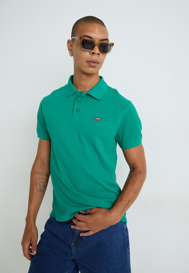 Polo verde in cotone, con colletto, chiusura a tre bottoni e un piccolo logo rosso Levi's. Indossato con jeans blu.