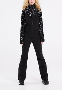 Čierne lyžovanie overall s predným zipsom, navrhnuté s nastaviteľnými popruhmi. Pod ním je upnutý dlhý rukáv top s bielym kvetinovým vzorom.