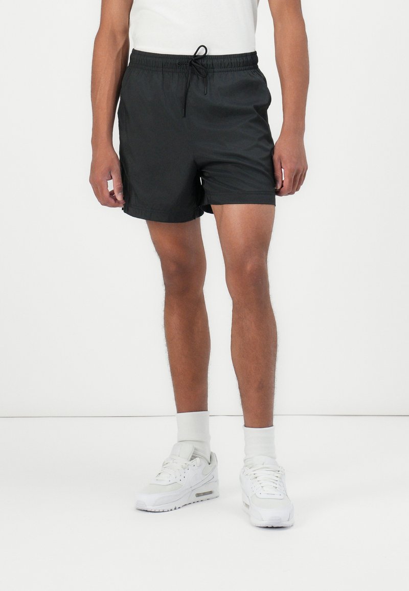 Svart svømmeshorts med elastisk midjebånd og justerbar snor, laget av et glatt og lett stoff. Brukt med hvite sneakers og sokker.