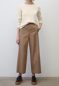 Haut beige côtelé à manches longues associé à un pantalon marron ample arrivant à la cheville. Chaussures noires à semelle épaisse complètent la tenue.