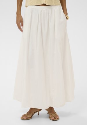 Femme portant une longue jupe blanche fluide avec une taille élastique et des sandales à talons beige, debout devant un fond uni.