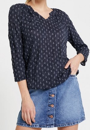 Femme portant un chemisier bleu marine avec motif d'ancre blanche et une jupe en jean bleue avec des boutons à l'avant, main reposant dans la poche de la jupe.